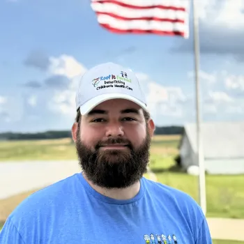 Bearded man in a Roof It Forward shirt and hat stands outside with a waving American flag and field background.