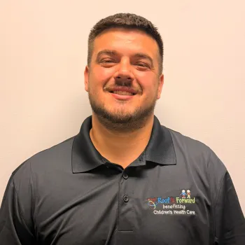 Smiling man wearing a black polo shirt with a colorful Roof It Forward charity logo on beige background