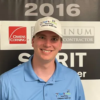 Smiling man in blue shirt and white hat with Children’s Health Care logos in front of a 2016 Owens Corning award banner.