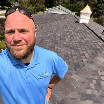 Man in blue shirt taking a selfie on a newly shingled rooftop with greenery and houses in the background.