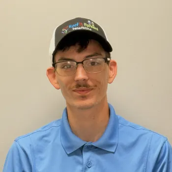 Young man wearing glasses and a Roof It Forward cap and blue polo shirt with charity logo on plain background