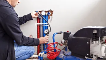 Technician checking water pressure gauges on pipes connected to a blue air compressor in a wall cavity.