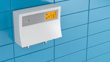 White digital thermostat mounted on blue tiled wall displaying temperature of 47.5 degrees Celsius.
