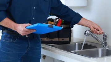 Man in blue shirt checking water flow from kitchen sink faucet with clipboard in hand and tools nearby.