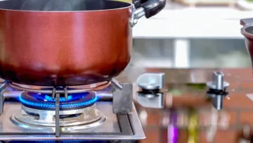 Red pot steaming on a gas stove burner with blue flame, next to a second pot on a reflective kitchen countertop.
