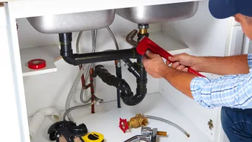 Plumber using a red wrench to fix black sink pipes under kitchen cabinet with tools on white shelf