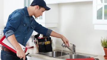 Plumber inspecting kitchen sink faucet with toolbox and wrench on countertop in modern white kitchen