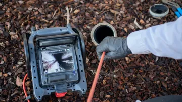 Plumber using a sewer camera to inspect a pipe underground surrounded by wood chips outdoors.