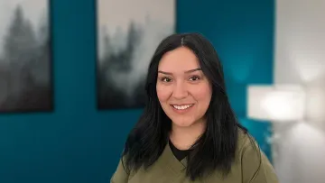 Smiling woman with long dark hair wearing olive green scrubs in a modern, softly lit room with blue walls.