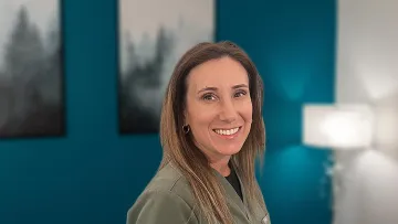 Smiling woman with long hair wearing a green medical scrub top against teal wall with blurred artwork and lamp.
