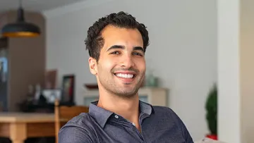 Smiling young man in a dark shirt sitting on a couch in a cozy, modern living room with warm lighting.