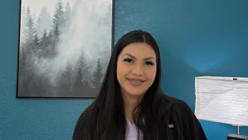 Smiling young woman with long dark hair and braces sits in blue room with forest painting and white lamp.