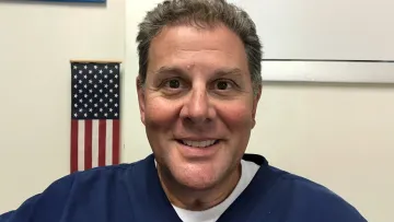 Smiling middle-aged man in blue scrub top stands in front of an American flag indoors.