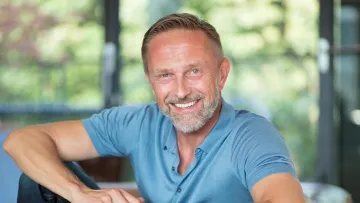 Smiling man sitting on a couch, dressed in a blue polo shirt, with natural light and greenery in the background.