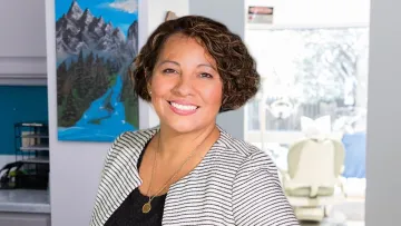 Smiling woman in a stylish outfit stands in a modern dental office with mountains painting.