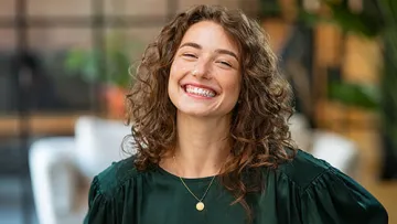 Smiling woman with curly hair wearing a dark green blouse and gold necklace in a cozy indoor setting
