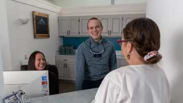 Smiling staff at a reception desk welcoming a visitor in a bright office setting.