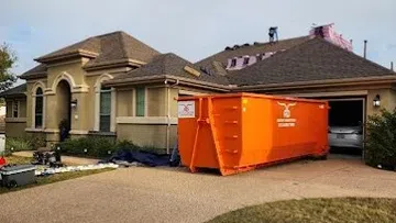 Residential house with roof under repair and large orange dumpster in driveway for construction debris removal.