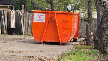 Bright orange Austin Dumpsters container on a driveway near trees and wooden fence in a residential area.