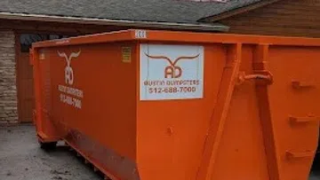 Large orange dumpster with Austin Dumpsters logo and contact number parked next to a building under cloudy sky.