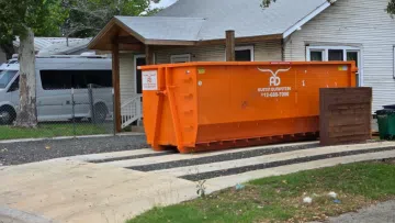 Large orange dumpster from Austin Dumpsters placed on a concrete pad in front of a white house with trees.
