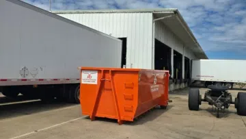 Orange dumpster container outside a warehouse with parked trailers under a partly cloudy sky.