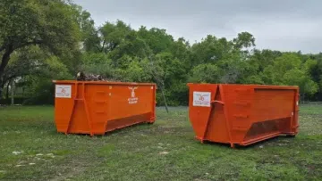 Two large orange construction dumpsters on grassy area with trees and cloudy sky in background