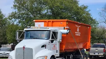 White semi truck hauling a large orange dumpster on a dirt road with trees and vehicles in the background