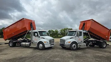 Two white dump trucks with raised orange containers parked on gravel under a cloudy sky with trees in the background