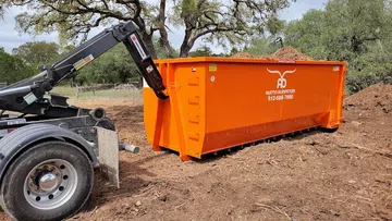 Orange Austin Dumpsters roll-off container loaded with debris on dirt ground with trees in background.