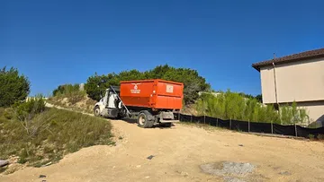 Orange dump truck on dirt road near hillside vegetation and building under clear blue sky.