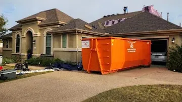 House with roof construction work in progress and large orange dumpster in driveway during daytime.