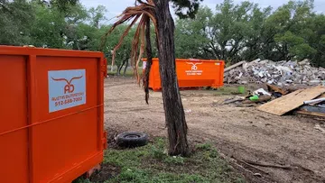 Two bright orange Austin Dumpsters at a debris cleanup site with trees and rubble in the background