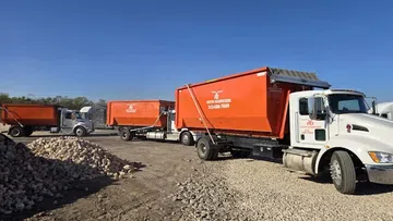 Orange dumpster trucks parked on gravel with blue sky at a construction or waste management site.