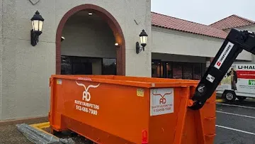 Large orange dumpster container with company logo and phone number outside beige building with tiled roof on wet pavement.