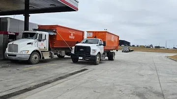 Two white trucks with orange trailers parked at a gas station on a cloudy day in a rural area