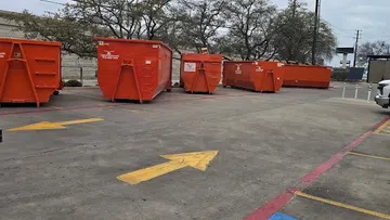 Orange dumpsters aligned in a parking lot with yellow arrows painted on the pavement and parked white trucks.