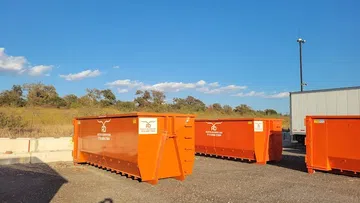 Three large orange industrial dumpsters on gravel under a clear blue sky near trees and a white building.