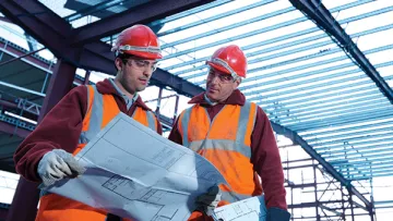 Two construction workers in safety vests and helmets reviewing blueprints inside a steel frame building under construction.
