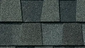 Large light blue house with gray shingle roof and front porch under a partly cloudy sky