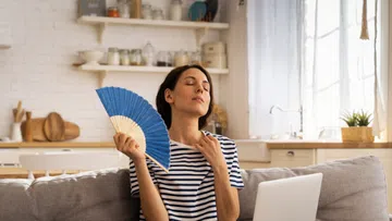 Woman sitting cross-legged on couch using a blue handheld fan to cool off with laptop on her lap in bright room
