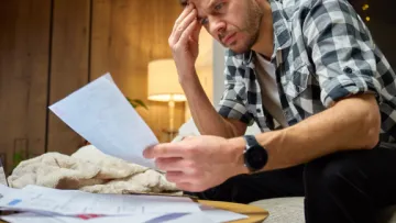 Young man in plaid shirt reviewing bills with a stressed expression in a cozy living room setting.