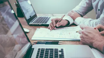 Two professionals collaborate over documents with laptops open on a wooden desk in a modern office setting.