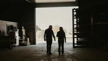 Silhouettes of two people walking and talking inside a warehouse with open door to bright exterior