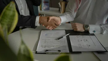 Two business professionals shaking hands over signed contract documents on a white desk with a pen.
