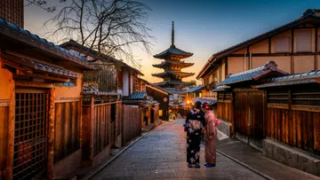 Traditional Japanese street at sunset with wooden houses, a pagoda, and two women in kimonos walking.