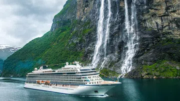 Large white cruise ship sailing near a towering green cliff with multiple waterfalls and cloudy sky.