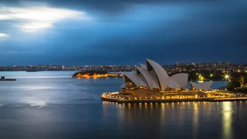 Sydney Opera House illuminated at night with city skyline and harbor waters under a dramatic sky.