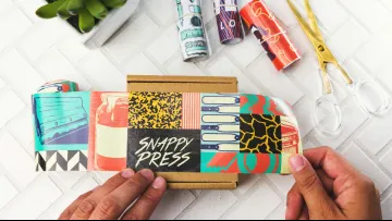 Person holding colorful Snappy Press printed wrap with craft scissors and succulent on white tile surface.