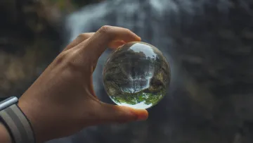 Hand holding a glass sphere reflecting an inverted waterfall and surrounding greenery in nature.